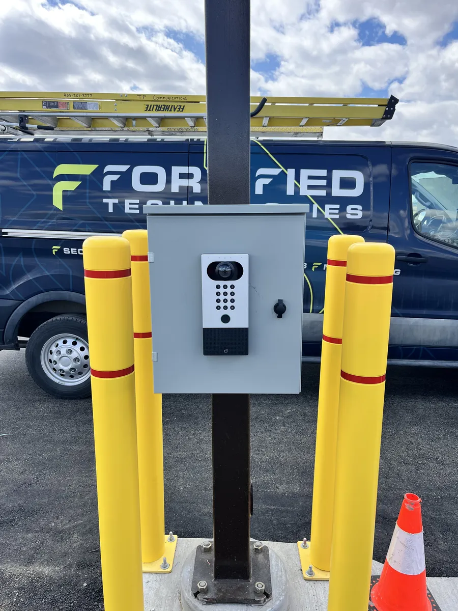 Video intercom and keypad access control unit mounted on a bollard-protected post beside a Fortified Technologies van