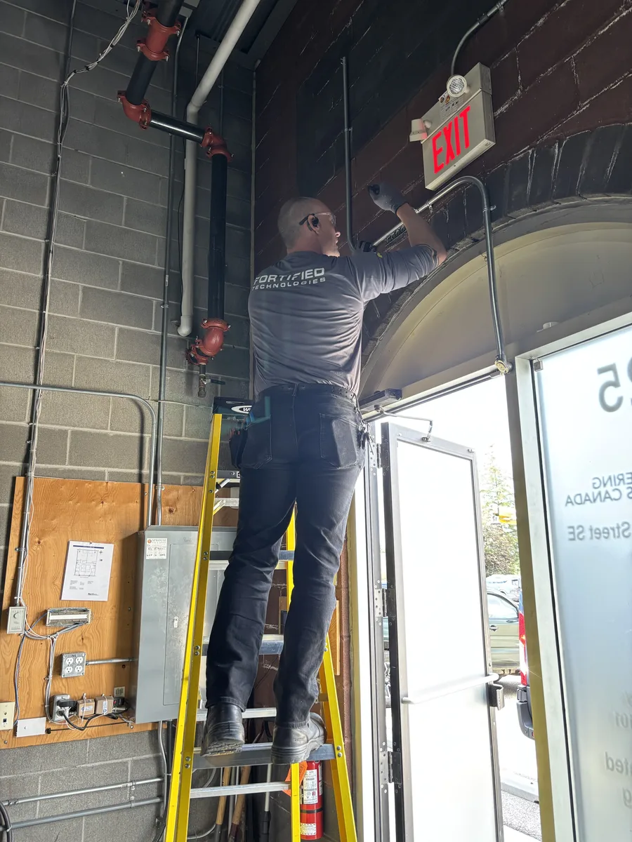 Fortified Technologies technician on a ladder installing security equipment above a commercial exit door