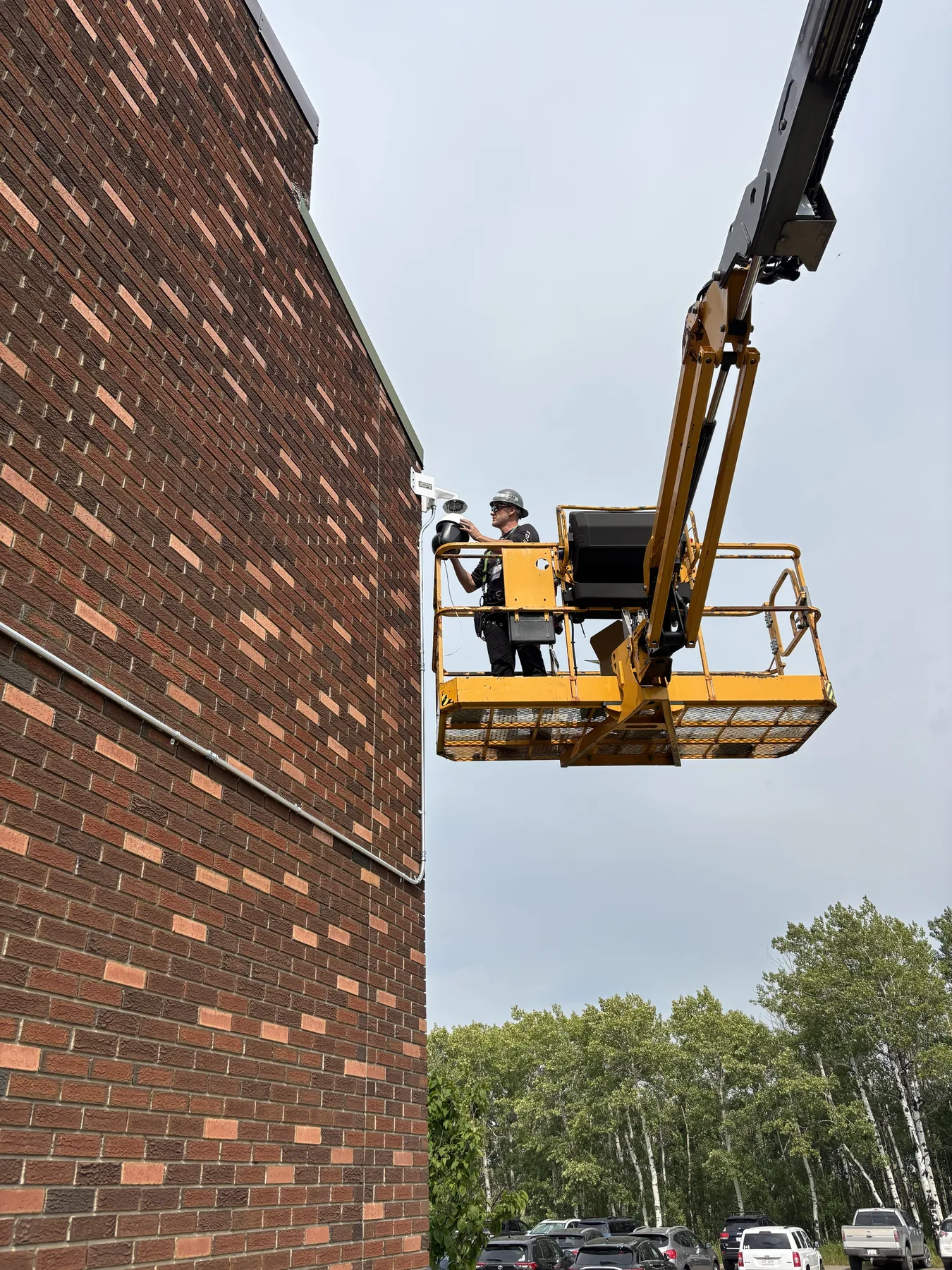 Fortified Technologies technician installing a security camera from an elevated work platform beside a brick building