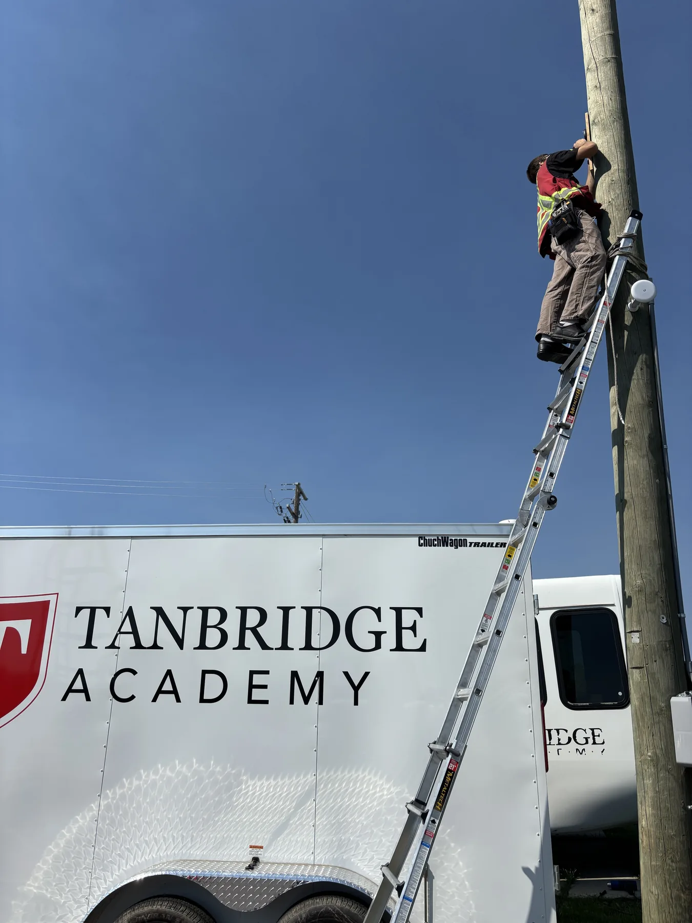 Fortified Technologies technician installing security equipment on a utility pole beside a Tanbridge Academy trailer